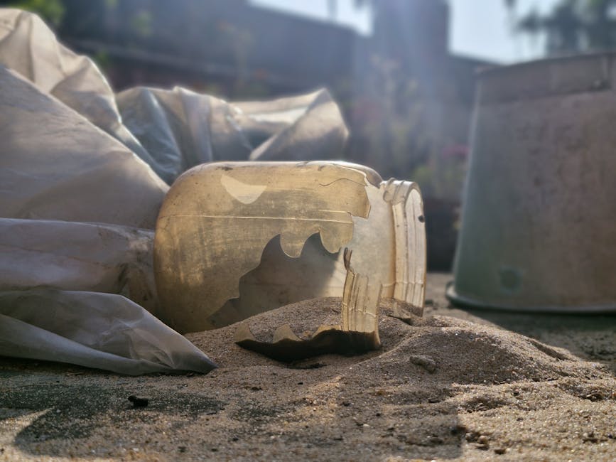 A close-up view of a broken glass jar with a jagged, uneven edge, lying on a sandy surface outdoors. The jar appears to be discarded, with fragments broken off and scattered nearby. Surrounding objects include a partially visible large plastic bag or wrapping material, and a metallic container or bucket in the background, both positioned on the sandy ground. The scene is captured in natural light, with a faint background of blurred outdoor structures and fencing, indicating a home renovation or cleaning process. The presence of debris and materials suggests packing or waste removal related to house relocation or renovation activities, with the broken glass highlighting the need for careful handling and disposal during removals. The image is presented in a clear, detailed manner, suitable for illustrating services related to rubbish clearance, packing, or logistics involved in furniture transport or home moves, as offered by Man with Van Sands End.