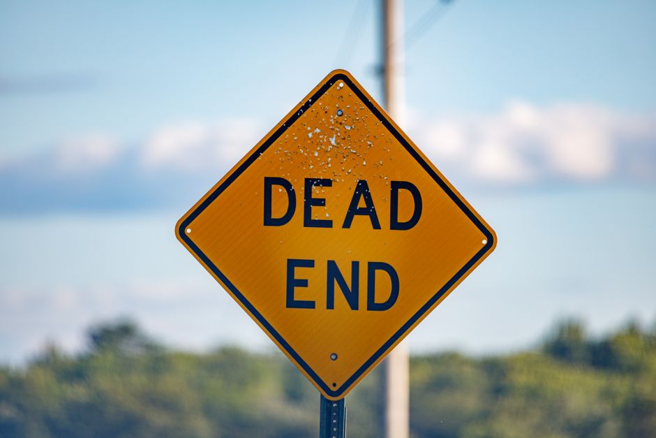 A yellow diamond-shaped road sign with black lettering reading 'DEAD END' is mounted on a metal pole outdoors. The sign appears weathered with visible water droplets on its surface. It is positioned near a boundary or potential obstacle, indicating a cul-de-sac or no-through road. In the background, there is a partly cloudy sky and blurred greenery, suggesting a suburban or semi-rural setting. This image captures the typical signage that may be encountered during home relocation or furniture transport processes, which Man with Van Sands End might consider when planning large moves or requiring road closures in Sands End. The outdoor environment and sign placement imply considerations relevant to moving logistics and route planning, especially in residential areas involved in packing and loading for house removals.