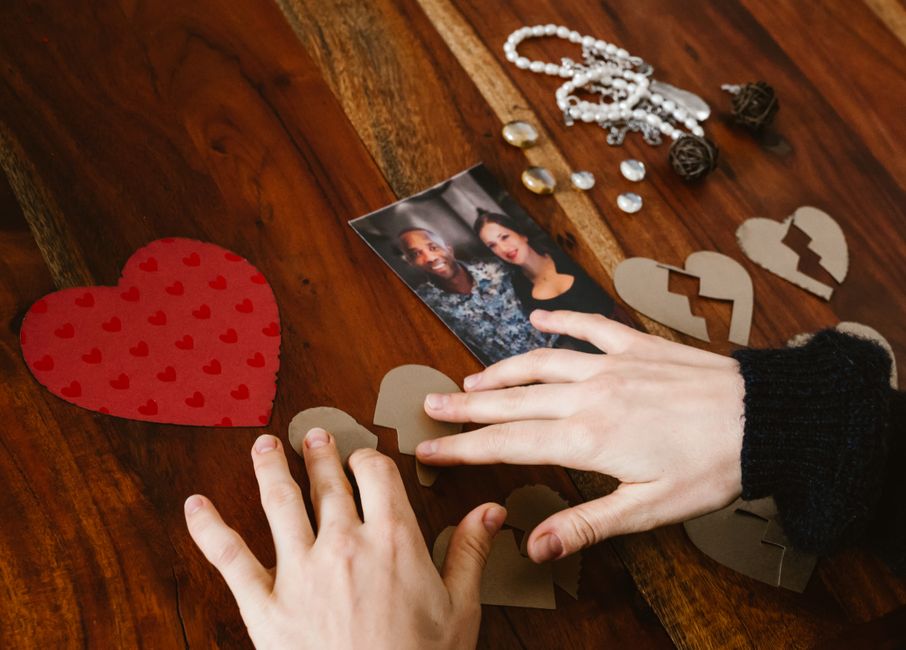 A pair of hands, one with a black knit sleeve, arranging torn cardboard heart-shaped pieces on a wooden table, alongside a printed photograph of two smiling women, a string of white pearls, decorative dark brown balls, and small loose gemstones. The scene appears to be set indoors with natural or warm lighting. The cardboard hearts include one large red heart with smaller red hearts printed on it, and several smaller, plain cardboard hearts, some of which are being positioned for a craft or symbolic activity related to home relocation or moving logistics, as associated with Man with Van Sands End's removals services in the context of packing and moving.