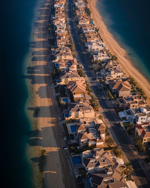 An aerial view of a residential area along a coastline showing rows of modern houses with tiled roofs, situated on a narrow street running parallel to the beach. The sidewalk and pavement are visible, with parked cars lining the street. The beach features golden sand extending alongside the calm, blue ocean. The scene is bathed in warm sunlight, highlighting the architectural details of the homes and the shadows cast by the houses and nearby vegetation. This image captures a coastal home neighbourhood, relevant to house removals and relocation services such as those offered by Man with Van Sands End, focused on facilitating home moves and furniture transport within the Fulham Reach and Sands End area.