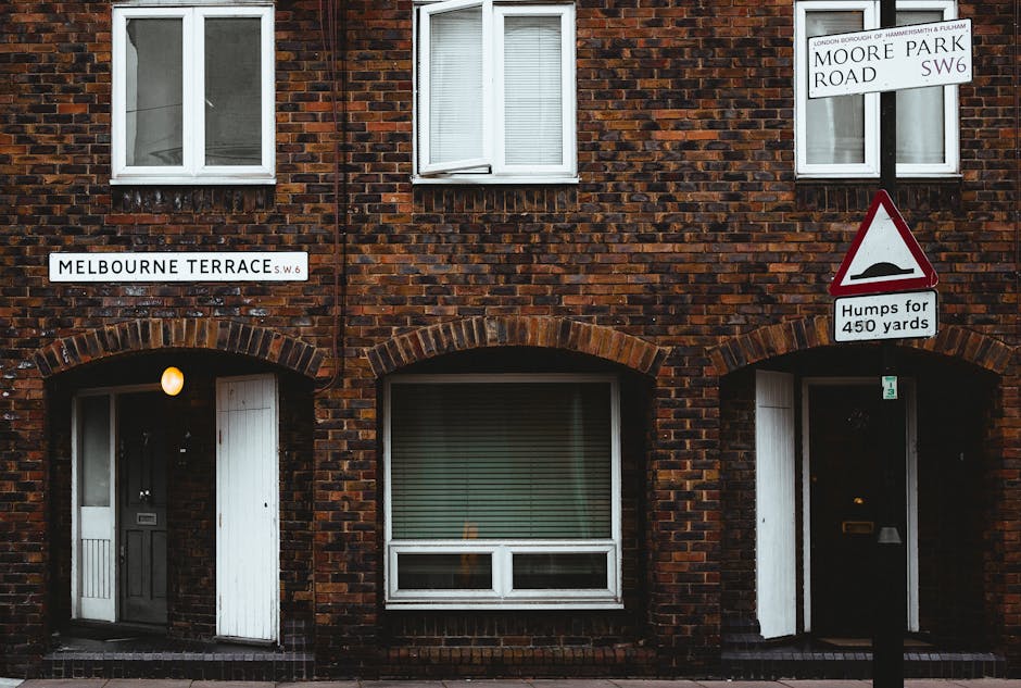 Photograph of a brick residential building at the corner of Melbourne Terrace and Moore Park Road in Hammersmith & Fulham, showing two white-framed windows on the upper floor and two doors beneath with arched brickwork above. One door, on the left, has a small frosted glass panel, a black door handle, and a light above it, illuminating the doorway. The other door, on the right, is dark with a mail slot. A white street sign reading 'Melbourne Terrace S.W.6' is mounted on the building's wall, and another sign on a pole indicates the area is part of Hammersmith & Fulham Council: Parking Permits for Moves, SANDS END. Additionally, a triangular warning sign with a hump symbol and text 'Humps for 450 yards' is visible, alerting drivers to road bumps. The scene suggests an urban setting suitable for home relocation or furniture transport activities involving removal services such as those provided by Man with Van Sands End, with visibility of a doorway, windows, and street signage supporting movement logistics.