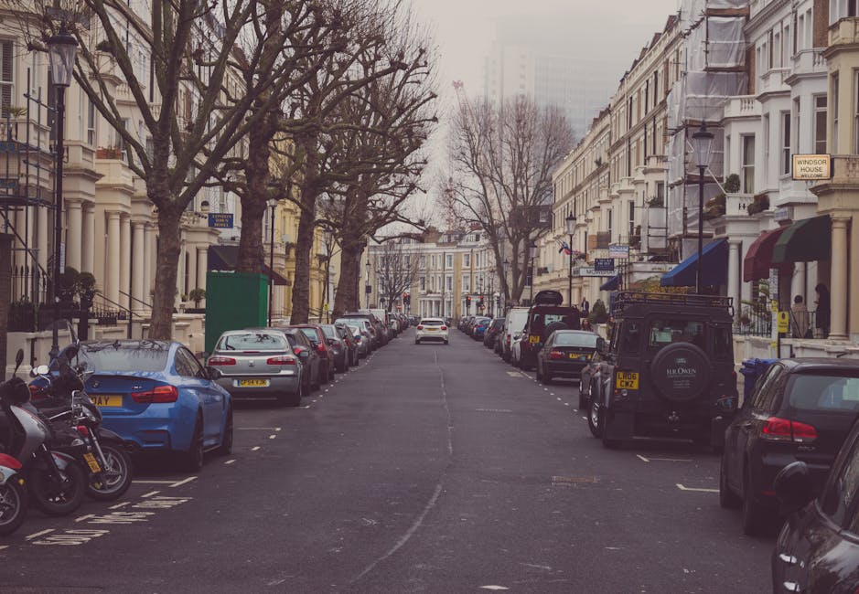 A residential street lined with parked cars and motorcycles on both sides, with multi-storey Victorian-style terraced houses featuring bay windows and decorative facades. Barren trees with twisted branches stretch overhead, suggesting late autumn or winter. In the background, a white van is being loaded or unloaded for house relocation, with visible moving blankets and cardboard boxes placed near the entrance of one property. The scene is captured during daylight with overcast sky lighting. A green access box is positioned on the sidewalk, and street signs indicate parking regulations. The image illustrates the typical setting for a house removal service in London, with [COMPANY_NAME] preparing furniture, boxes, and moving equipment as part of the home relocation process, within a controlled urban environment consistent with street parking and moving logistics in Hammersmith & Fulham.
