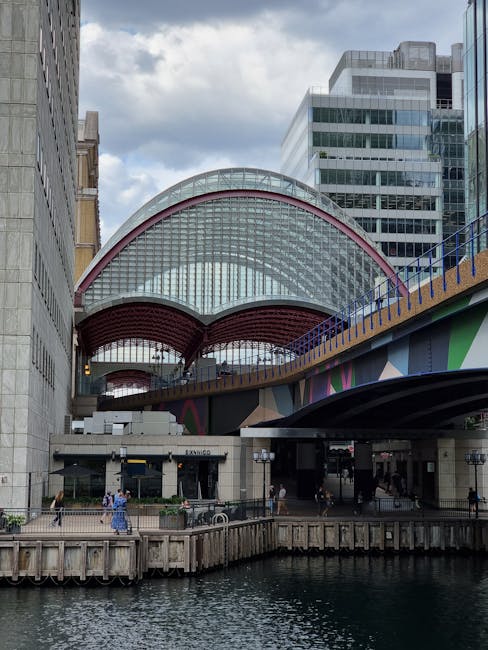 A modern urban scene at Imperial Wharf in Sands End featuring a contemporary glass and steel station building with a large, semi-circular, red-trimmed roof structure. The station is elevated, with a colorful painted bridge or walkway underneath, supported by dark steel columns. In the foreground, a waterway with a wooden quay runs alongside the station, with pedestrians walking along the pavement and waiting near the entrance. The surrounding area includes high-rise buildings with reflective glass facades, and the sky above is partly cloudy. The image depicts a busy, mixed-use environment suitable for home relocation or moving services, with visible infrastructure for transportation and pedestrian access, typical of a location serviced by professional removals companies like Man with Van Sands End.
