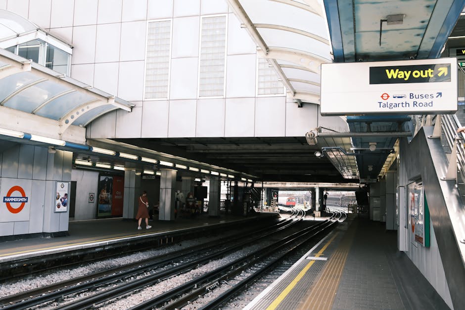 Exterior view of Hammersmith station with multiple train tracks and platform edge visible in the foreground. The station features a modern, metallic cladding with white panels and clear signage indicating directions for buses to Talgarth Road and a pathway labeled 'Way out' with an arrow. A staircase with metal railings ascends on the right side of the image, and a woman is seen walking along the platform wearing casual clothing. Inside the station, the ceiling is supported by white, curved structures, and there are large windows letting in natural light. This setting illustrates a typical urban transportation hub in London, emphasizing the connectivity relevant to home relocation and moving services, such as those provided by Man with Van Sands End, which involves careful planning of luggage and equipment transportation, including loading and unloading processes near the station infrastructure.