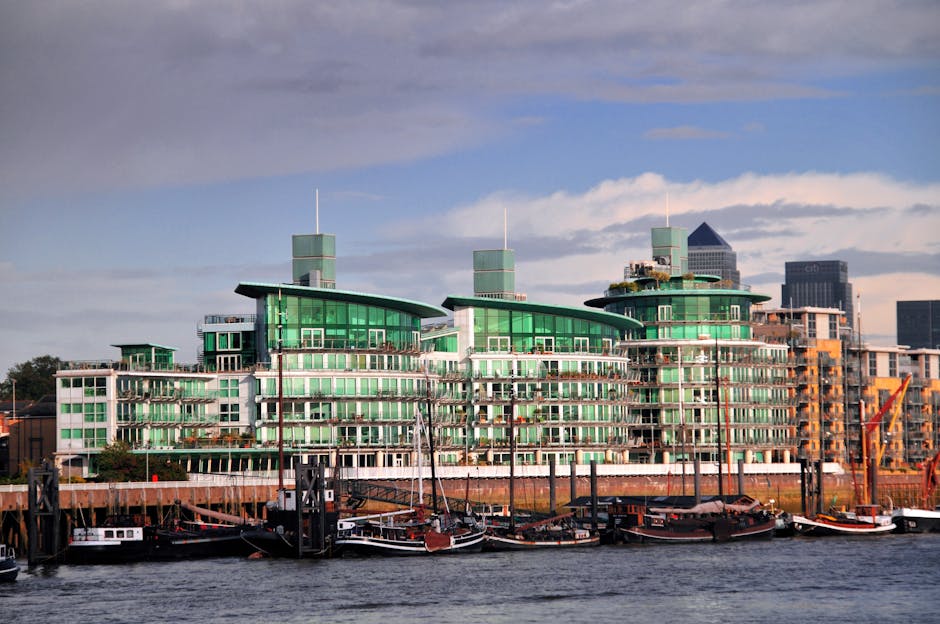 A modern urban scene at Imperial Wharf in Sands End featuring a contemporary glass and steel station building with a large, semi-circular, red-trimmed roof structure. The station is elevated, with a colorful painted bridge or walkway underneath, supported by dark steel columns. In the foreground, a waterway with a wooden quay runs alongside the station, with pedestrians walking along the pavement and waiting near the entrance. The surrounding area includes high-rise buildings with reflective glass facades, and the sky above is partly cloudy. The image depicts a busy, mixed-use environment suitable for home relocation or moving services, with visible infrastructure for transportation and pedestrian access, typical of a location serviced by professional removals companies like Man with Van Sands End.