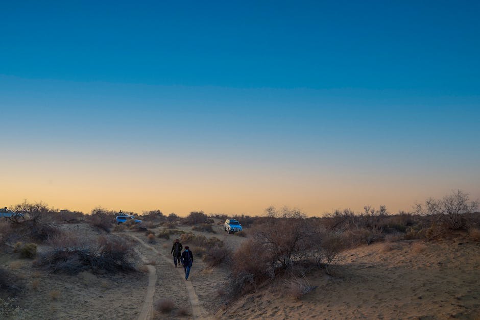 Two individuals walking along a narrow dirt track through a desert landscape during late afternoon or early evening, with sparse bushes and small trees on either side. In the background, a white moving van with blue accents is parked on a slightly elevated area, partly covered with a protective fabric or blanket, indicating a home relocation or furniture transport process. The scene is illuminated by natural light, with a deep blue sky transitioning to a warm orange near the horizon, suggesting the end of the day. The setting appears remote and open, with no nearby buildings, emphasizing the outdoor environment typical for loading or unloading items during a house move. The presence of moving vehicles and the rural terrain subtly reflect the logistics involved in local or long-distance removals, consistent with services offered by Man with Van Sands End.