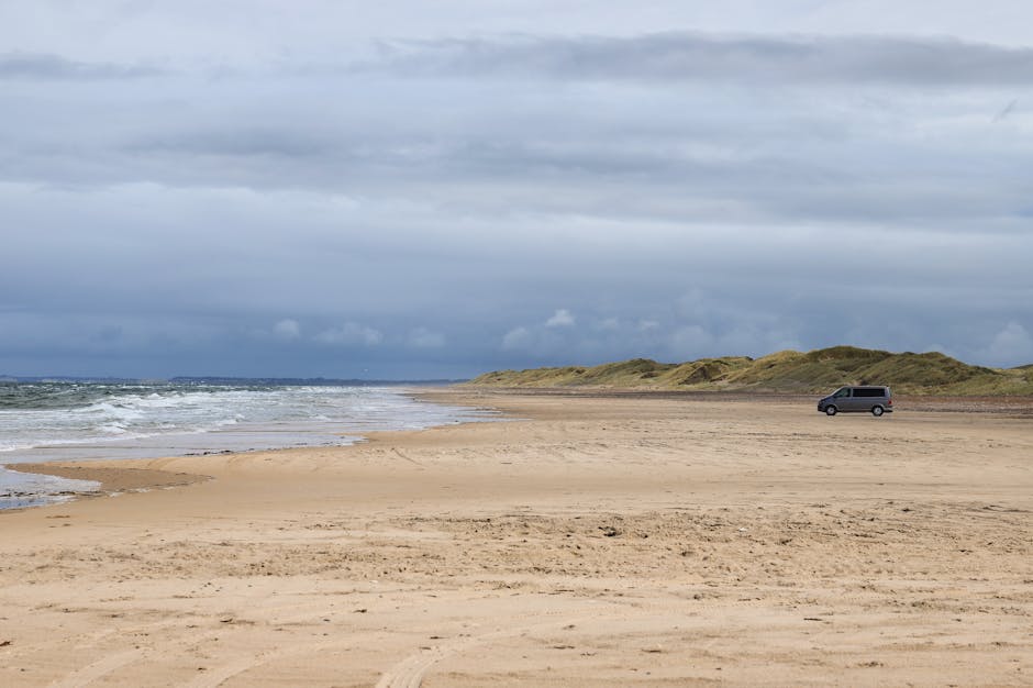 A grey van parked on a deserted sandy beach near the shoreline, with gentle waves rolling onto the shore. The beach is large and flat, with tire tracks visible in the sand, and bordered by grassy dunes in the background under a cloudy, overcast sky. The image appears to be taken during daytime, with diffuse natural light illuminating the scene. In the context of house removals or home relocation, this scene might represent a vehicle used for transport or a staging area for a moving operation, as seen by the presence of the van on the expansive beach. Man with Van Sands End could potentially use a similar vehicle for furniture transport and packing during same-day or last-minute moves, supported by the loaded and organized appearance of the van for moving services.