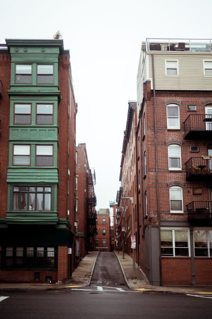 A narrow urban residential street during daylight lined with multi-storey brick buildings, one on the left painted in green with large windows and external black metal fire escapes, and another on the right in beige with balconies and a rooftop terrace. The street has a steep incline with a concrete driveway leading uphill, flanked by pavement on both sides, and is free of vehicles or pedestrians. The sky is overcast, and the scene depicts a typical location for home relocation or furniture transport, where careful loading and moving logistics are necessary, often involving professional removals services like those provided by Man with Van Sands End for house removals and relocation projects.