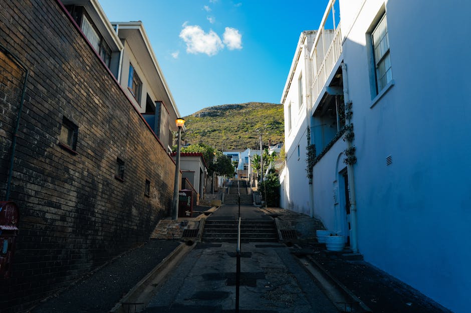 A steep urban street in Sands End with residential buildings on either side, including a white-painted building on the right with a visible balcony and window, and a darker brick building on the left. The street features stone steps and a central handrail leading up the incline toward hillside homes and greenery in the background. The sky is clear with some clouds, and a streetlamp is illuminated along the sidewalk. The scene depicts the challenging stairs and incline that may be encountered during house removals and furniture transport in the area. Man with Van Sands End, a removals company, may coordinate services to navigate this staircase for home relocation and packing and moving efforts.