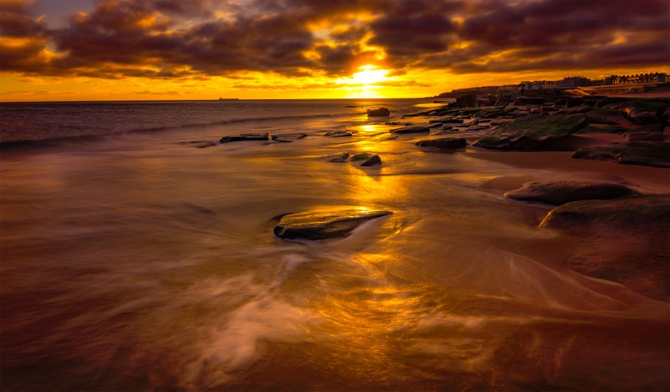 A silhouetted figure of a person stands on a sandy beach during sunset, with warm, golden light illuminating the sky and reflecting on the water. In the background, two other figures are visible at a