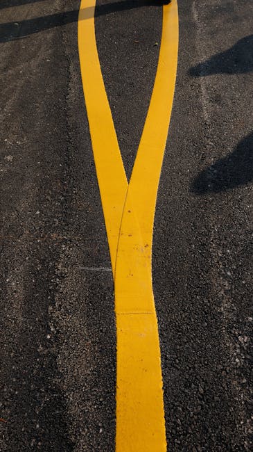 A close-up view of a section of tarmac pavement painted with bright yellow double lines, slightly curved and showing signs of wear and dirt. The lines are used for traffic regulation in residential or urban areas, such as in Sands End. The pavement surrounds a moving or parking area where furniture and boxes are likely being loaded onto a van, with the edge of a moving vehicle visible in the background. This scene is representative of home relocation activities, involving careful parking or street management during furniture transport and packing processes, as seen in professional removals services by Man with Van Sands End.
