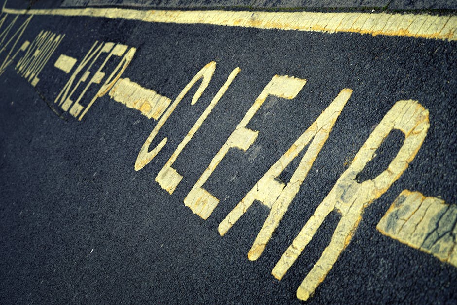 A close-up view of a section of tarmac pavement painted with bright yellow double lines, slightly curved and showing signs of wear and dirt. The lines are used for traffic regulation in residential or urban areas, such as in Sands End. The pavement surrounds a moving or parking area where furniture and boxes are likely being loaded onto a van, with the edge of a moving vehicle visible in the background. This scene is representative of home relocation activities, involving careful parking or street management during furniture transport and packing processes, as seen in professional removals services by Man with Van Sands End.
