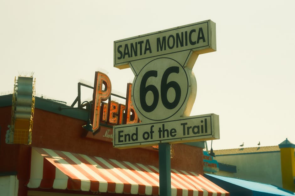 A detailed close-up view of a vintage Route 66 road sign mounted on a pole, indicating 'Santa Monica' at the top and 'End of the Trail' at the bottom, situated in a lively urban area. Behind the sign, part of a building with a red and white striped awning is visible, along with a brightly lit neon sign displaying the word 'PEP' in orange. The scene is captured during daylight with a clear sky, and some pigeons are perched on the rooftops. The image showcases elements related to travel and landmark destinations, subtly aligning with house removals and moving services by highlighting transportation and navigation themes associated with relocations. Man with Van Sands End often facilitates furniture transport and home relocation, emphasizing smooth logistics during moving processes.
